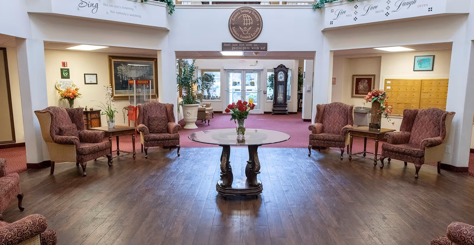 A spacious senior living facility lobby with wooden flooring and a central round table holding a vase of red and yellow flowers. The seating area includes several upholstered armchairs arranged around small side tables with plants and flowers. The background shows a grandfather clock, glass entrance doors, and mailboxes on the right side. Inspirational quotes are displayed on the walls above the seating area.
