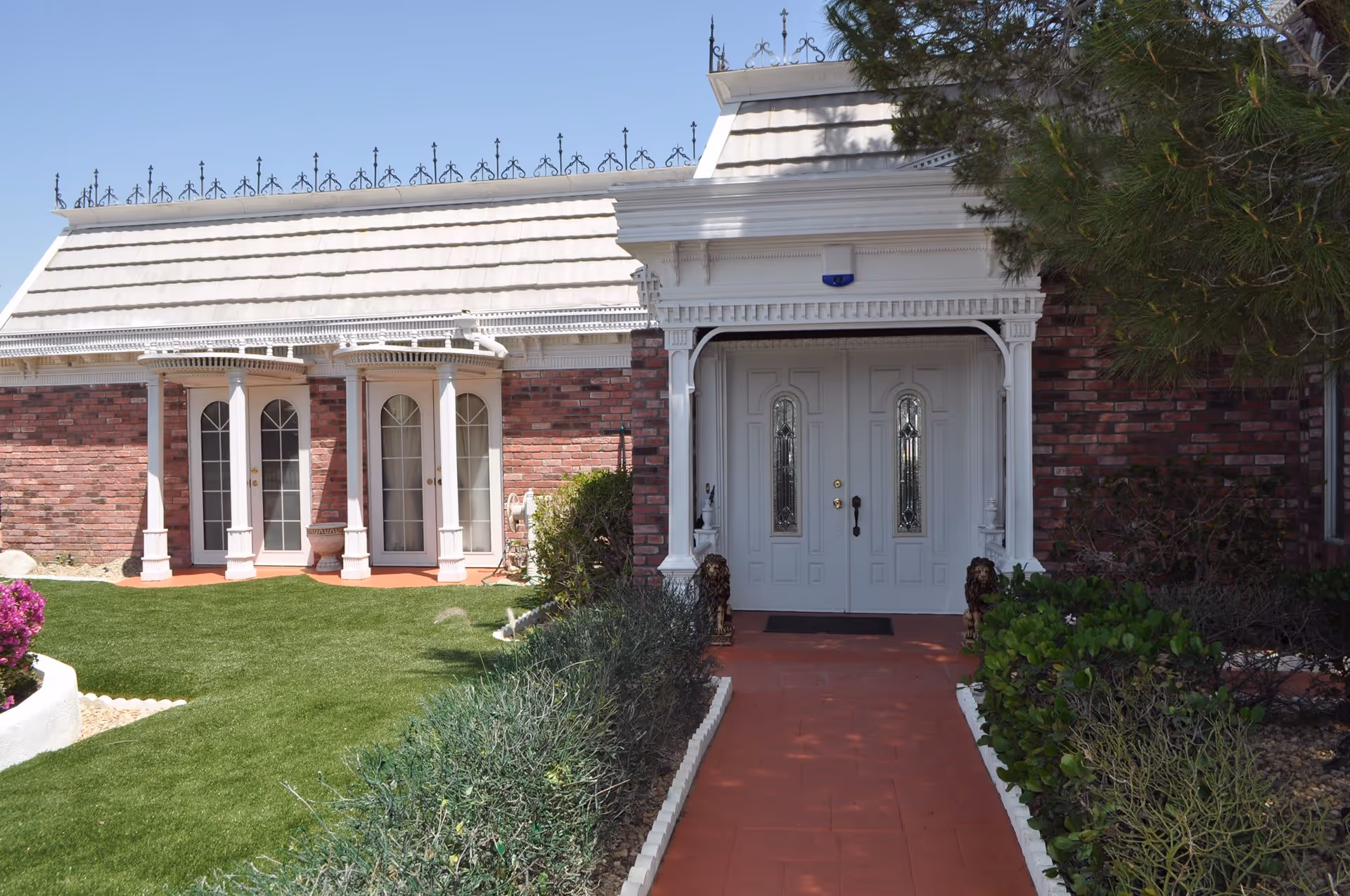 Front entrance of a brick building with white double doors featuring decorative glass panels, a white roof with ornamental ironwork, and a red brick pathway leading to the door. The entrance is flanked by bushes and small lion statues, with a well-maintained lawn and flowering plants nearby.