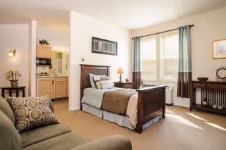 A bright and cozy bedroom in a senior living facility featuring a single wooden bed with pillows and a brown blanket. There is a nightstand with a lamp beside the bed, a cushioned armchair with patterned pillows, and a window with curtains letting in natural light. The room also includes a small table with decorative items and a clock, and a view into a kitchenette area.
