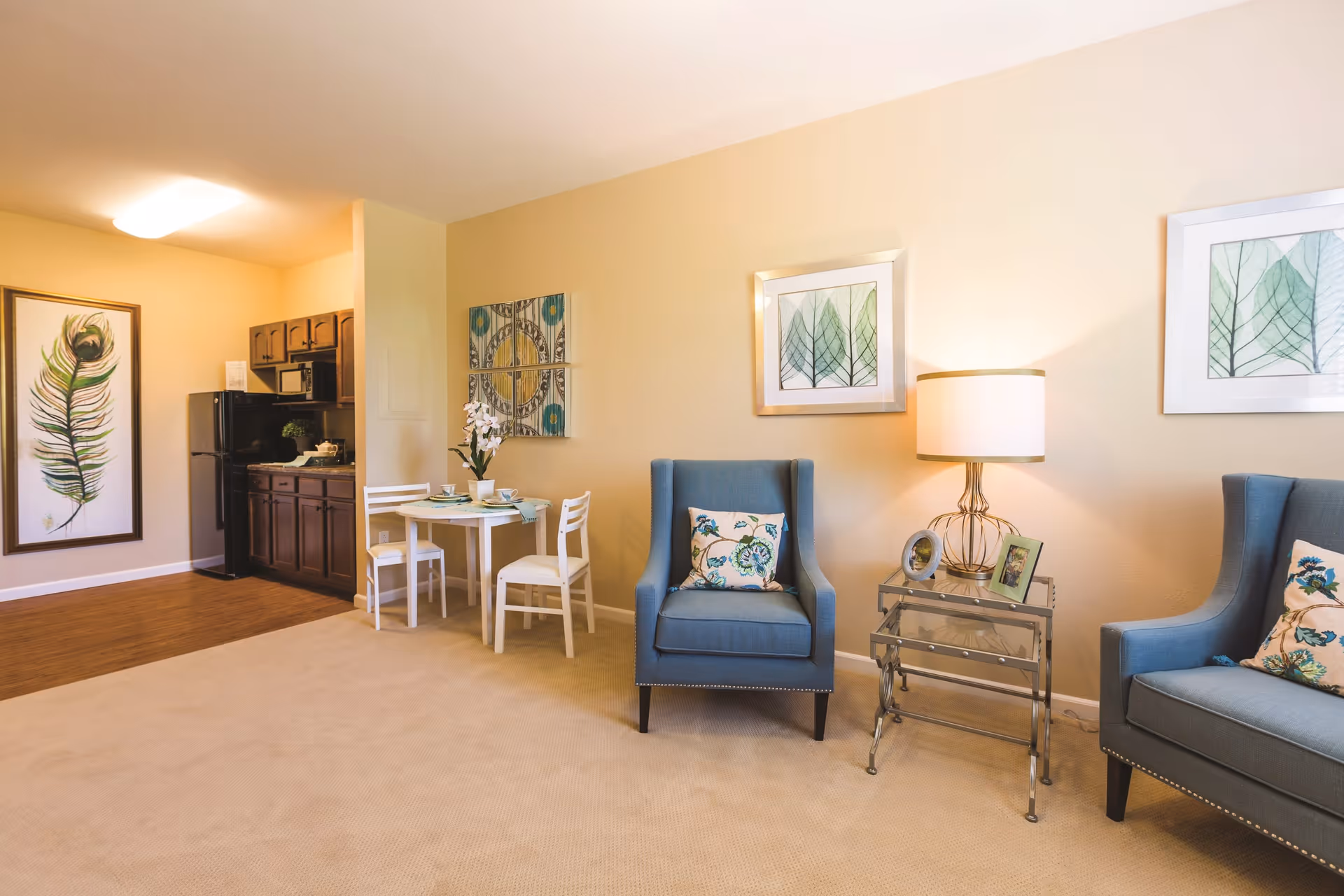 A cozy living area in an assisted living facility featuring two blue armchairs with floral cushions, a glass side table with a lamp and framed photos, a small round dining table with two white chairs, and a kitchenette with dark wood cabinets and a black refrigerator. The walls are decorated with framed artwork including leaves and a peacock feather.