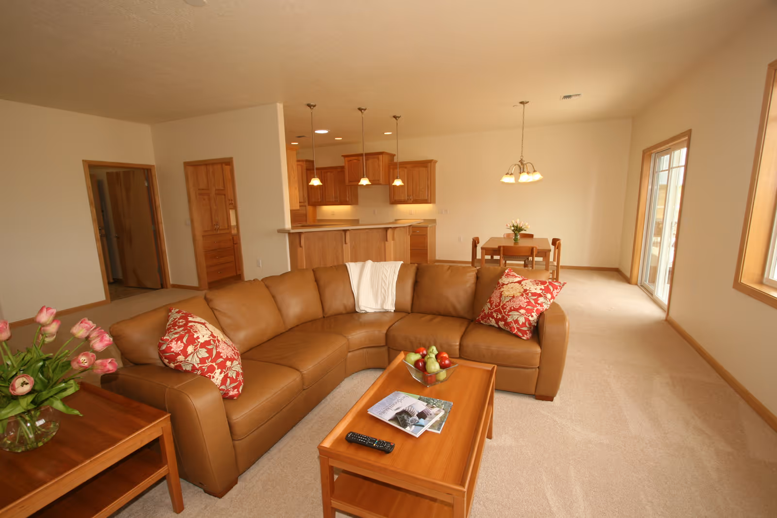 A spacious living room with a brown leather sectional sofa adorned with red and white floral pillows and a white throw blanket. In front of the sofa is a wooden coffee table with a bowl of fruit, a remote control, and magazines. To the left is a wooden side table with a vase of pink tulips. The room has beige carpet and cream-colored walls with wooden trim. In the background, there is an open kitchen with wooden cabinets and three pendant lights hanging over a breakfast bar. A small dining area with a wooden table and chairs is visible near large windows and a glass door letting in natural light.