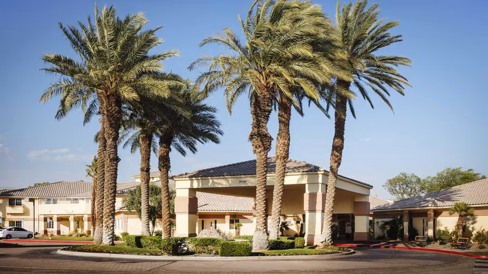 Exterior view of Atria Rancho Mirage senior living facility with tall palm trees in front, a covered entrance, and beige buildings with tiled roofs under a clear blue sky.