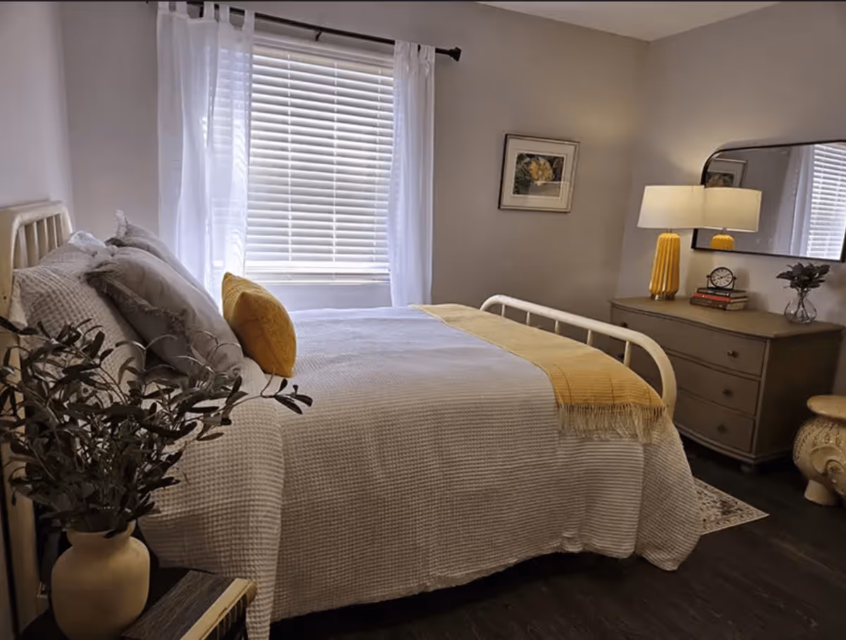 A cozy bedroom with a neatly made bed featuring white textured bedding and yellow accent pillows and throw. The room has a window with white blinds and sheer curtains, a dresser with a yellow lamp, clock, books, and a small plant, a framed floral picture on the wall, and a potted plant on a small table near the bed.