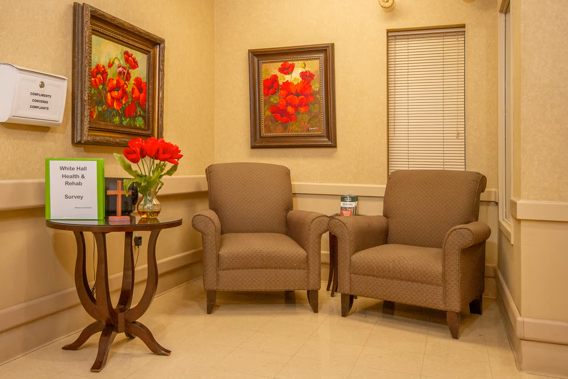 A cozy seating area with two brown upholstered armchairs placed side by side against a beige wall. Between the chairs is a small round wooden table holding a vase with red flowers, a small cross, and a green folder labeled 'White Hall Health & Rehab Survey'. Two framed paintings of red flowers hang on the wall above the chairs. A white box labeled 'Compliments Concerns Complaints' is mounted on the wall to the left.