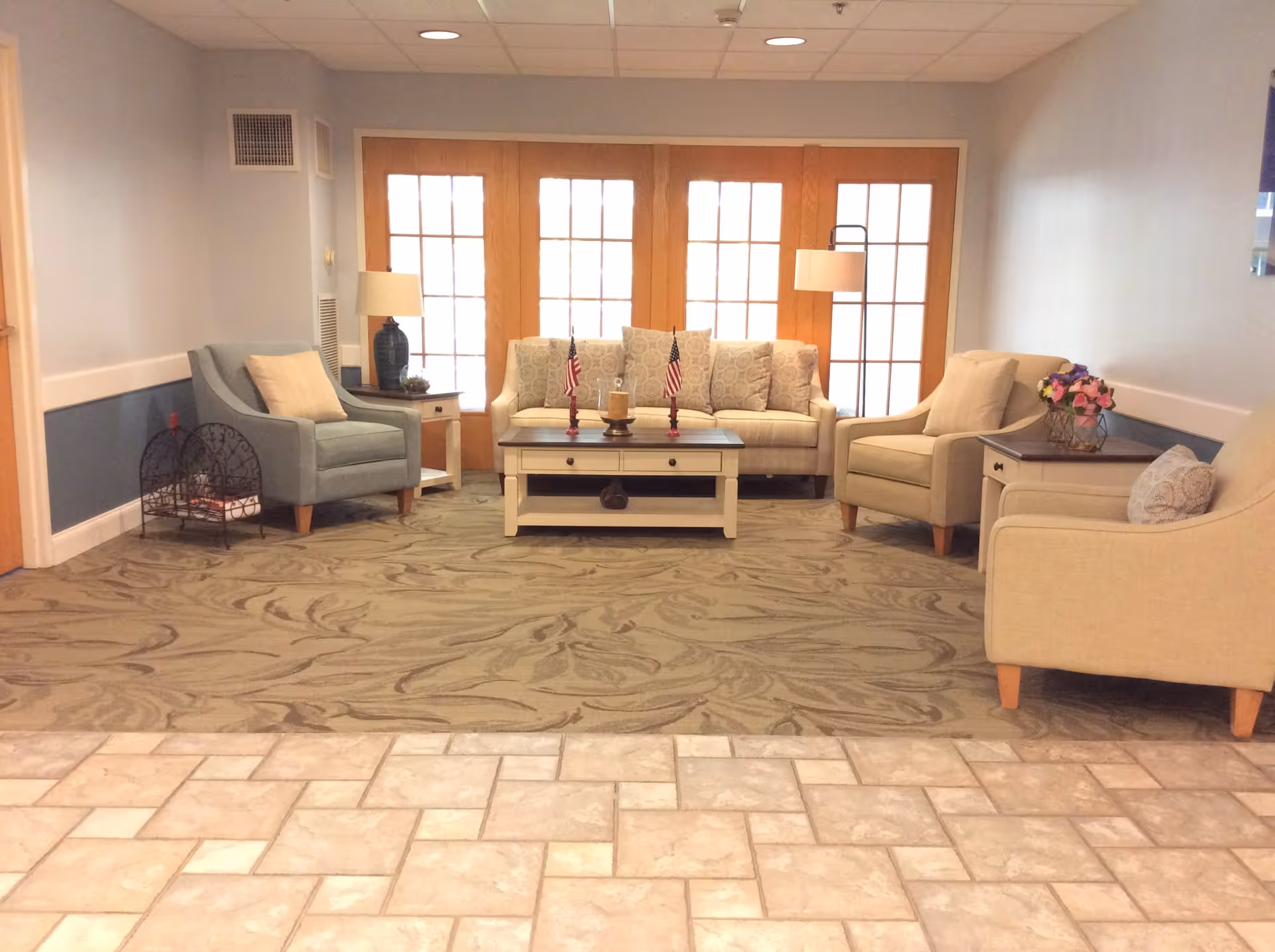 A cozy sitting area in The Cascades Assisted Living featuring a beige sofa with patterned cushions, two beige armchairs, and one blue armchair arranged around a white coffee table with two small American flags and a candle. The area is carpeted with a patterned rug, and there are two side tables with lamps and a flower arrangement. Behind the seating area are large wooden framed glass doors letting in natural light.