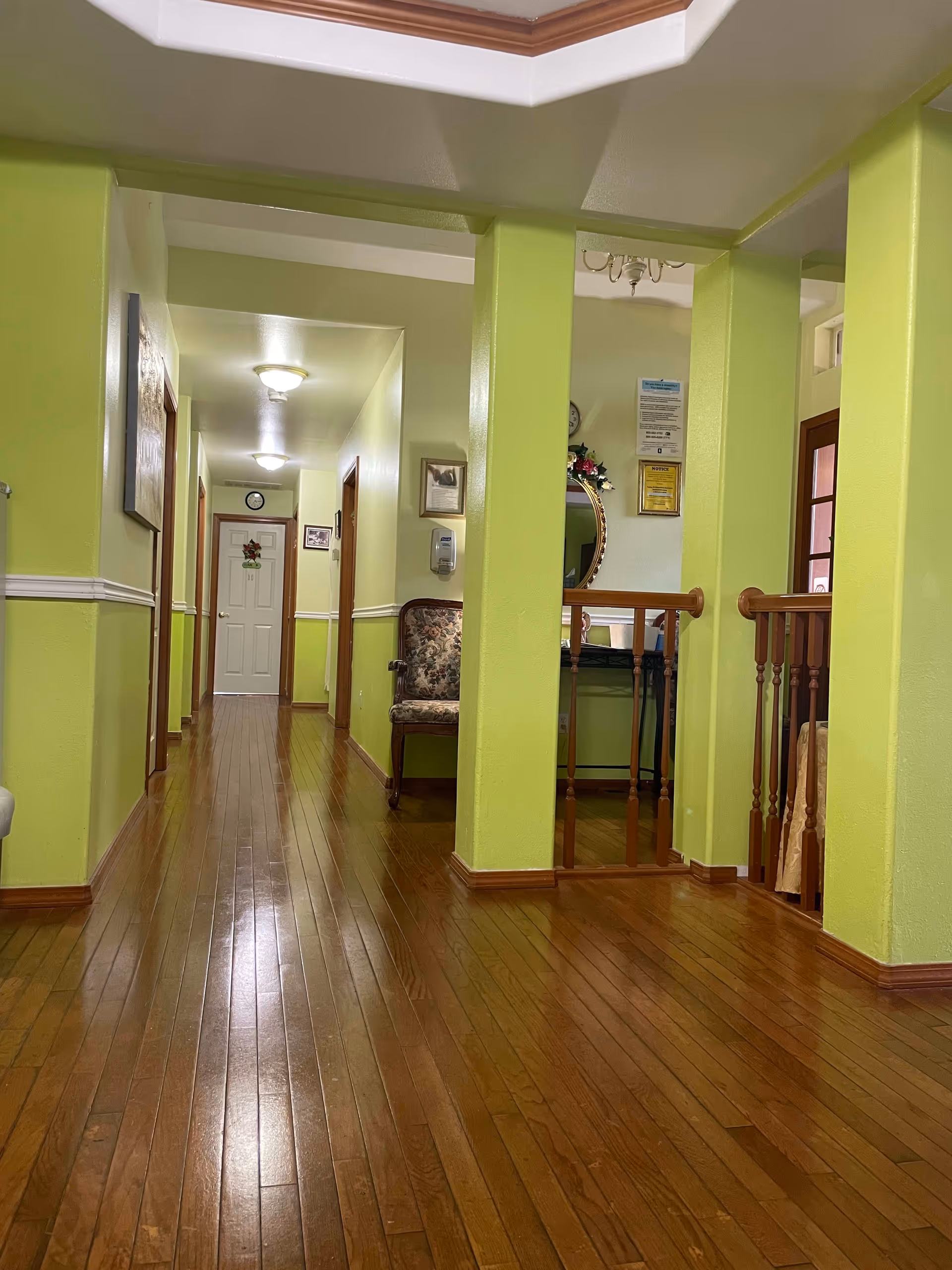 Interior hallway of a facility with light green walls and wooden flooring. The hallway has several doors along the sides and a white door at the end. There is a floral upholstered chair against the wall and a small table with a round mirror and some framed notices on the wall. The ceiling has recessed lighting and a decorative ceiling feature.