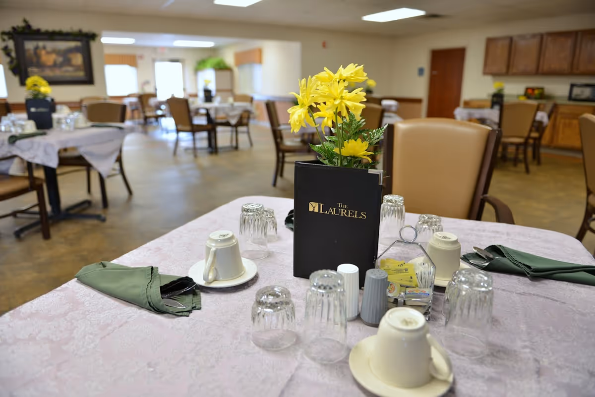 A dining room table set with cups, glasses, green napkins and a menu labeled "The Laurels" with a yellow flower centerpiece in a senior living facility dining area.
