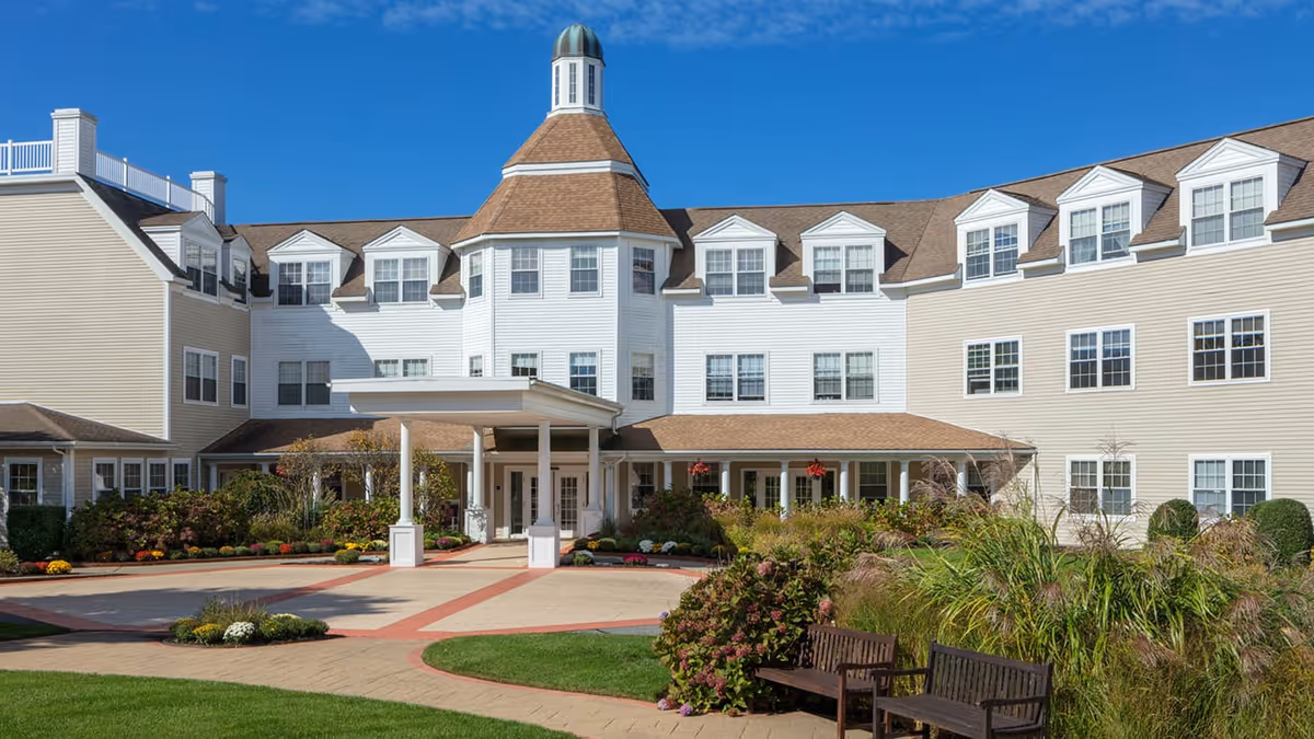 Front exterior view of a large senior living facility building with beige siding, multiple windows, a central tower with a cupola, a covered entrance, landscaped gardens, and benches on a sunny day with clear blue sky.