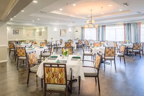 A bright and spacious dining room with multiple tables covered in white tablecloths, each set with green napkins, cups, and small flower arrangements. The room features large windows with curtains allowing natural light to fill the space, modern ceiling lights, and wooden chairs with patterned upholstery.
