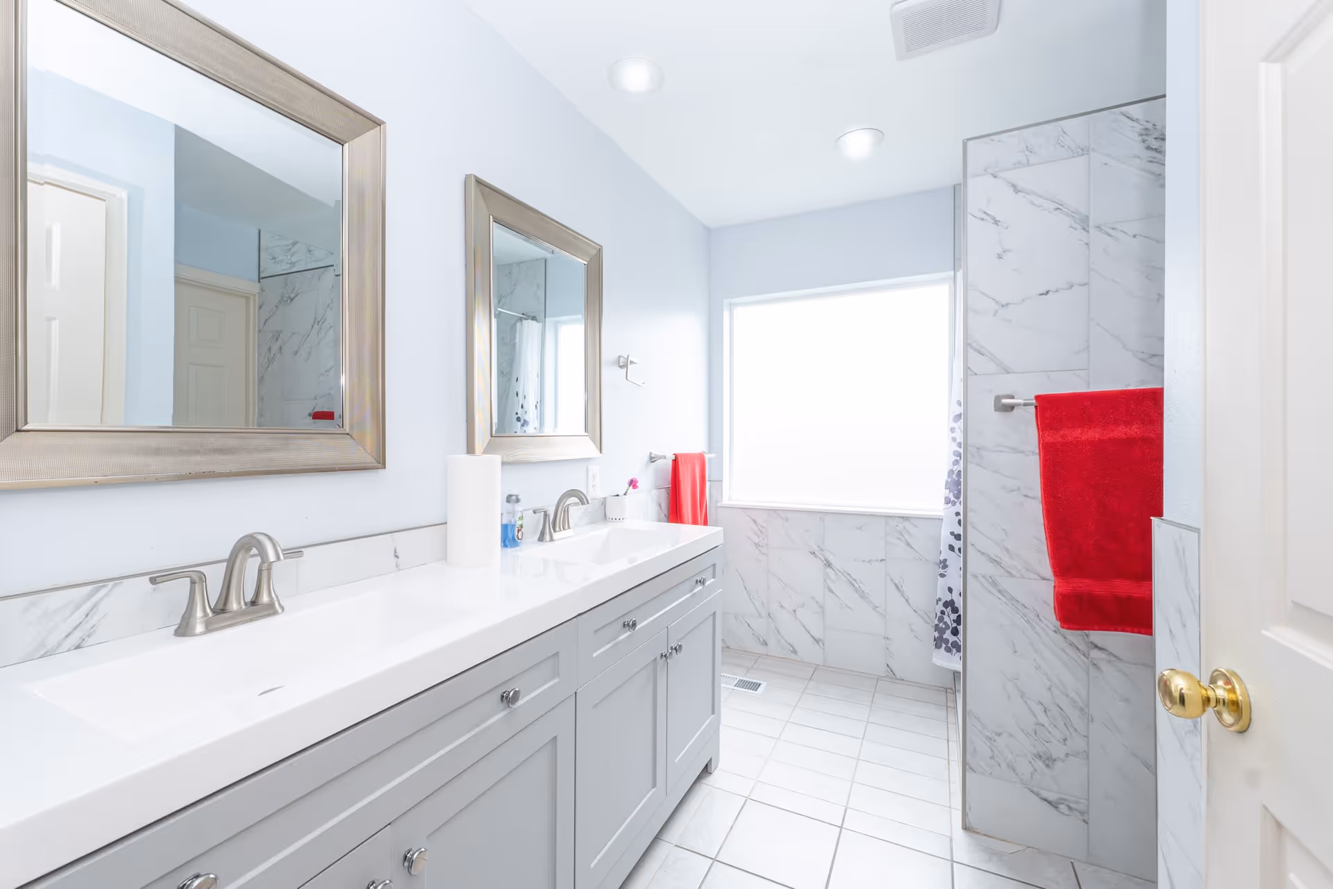 Bright bathroom with a double sink vanity featuring silver faucets and two large framed mirrors above. The walls are light blue, and the floor is tiled in white. There is a window letting in natural light, a shower area with marble-patterned tiles, and a red towel hanging on a towel rack.