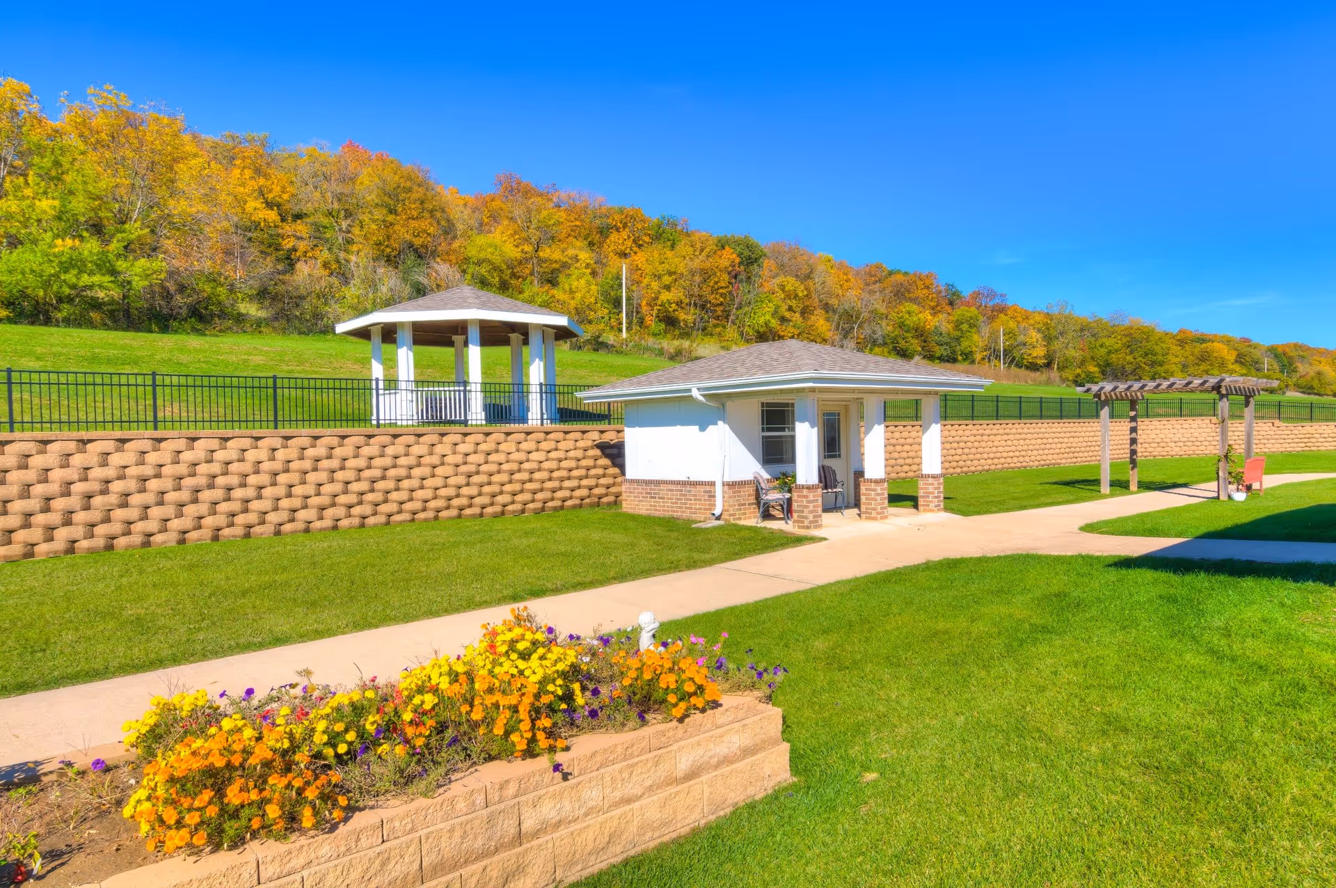 Outdoor area of River Valley Place of Fort Madison featuring a small white building with a covered porch, a gazebo on a raised grassy area behind a retaining wall, a pergola, a flower bed with colorful flowers, and a paved walkway under a clear blue sky.