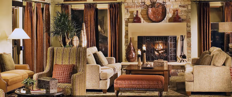 Cozy living room with beige sofas, a patterned armchair, a wooden coffee table with candles, and a stone fireplace with a fire burning. The room has brown curtains and warm lighting from lamps.