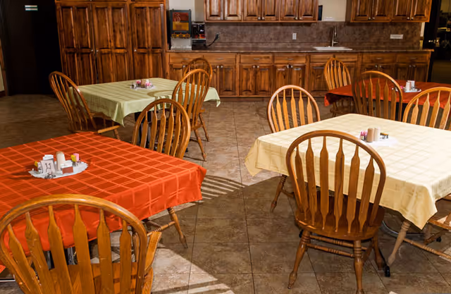 A dining room with several wooden tables covered with red, yellow, and green tablecloths. Each table has wooden chairs around it and small condiment holders on top. The background shows wooden cabinets and a countertop with a coffee machine.