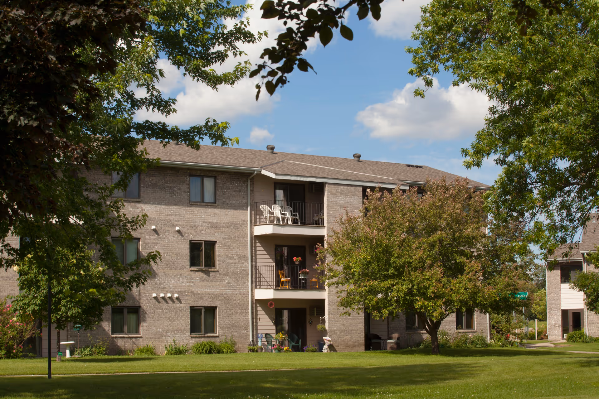 Exterior view of a three-story brick residential building with balconies, surrounded by green trees and a well-maintained lawn under a partly cloudy sky.