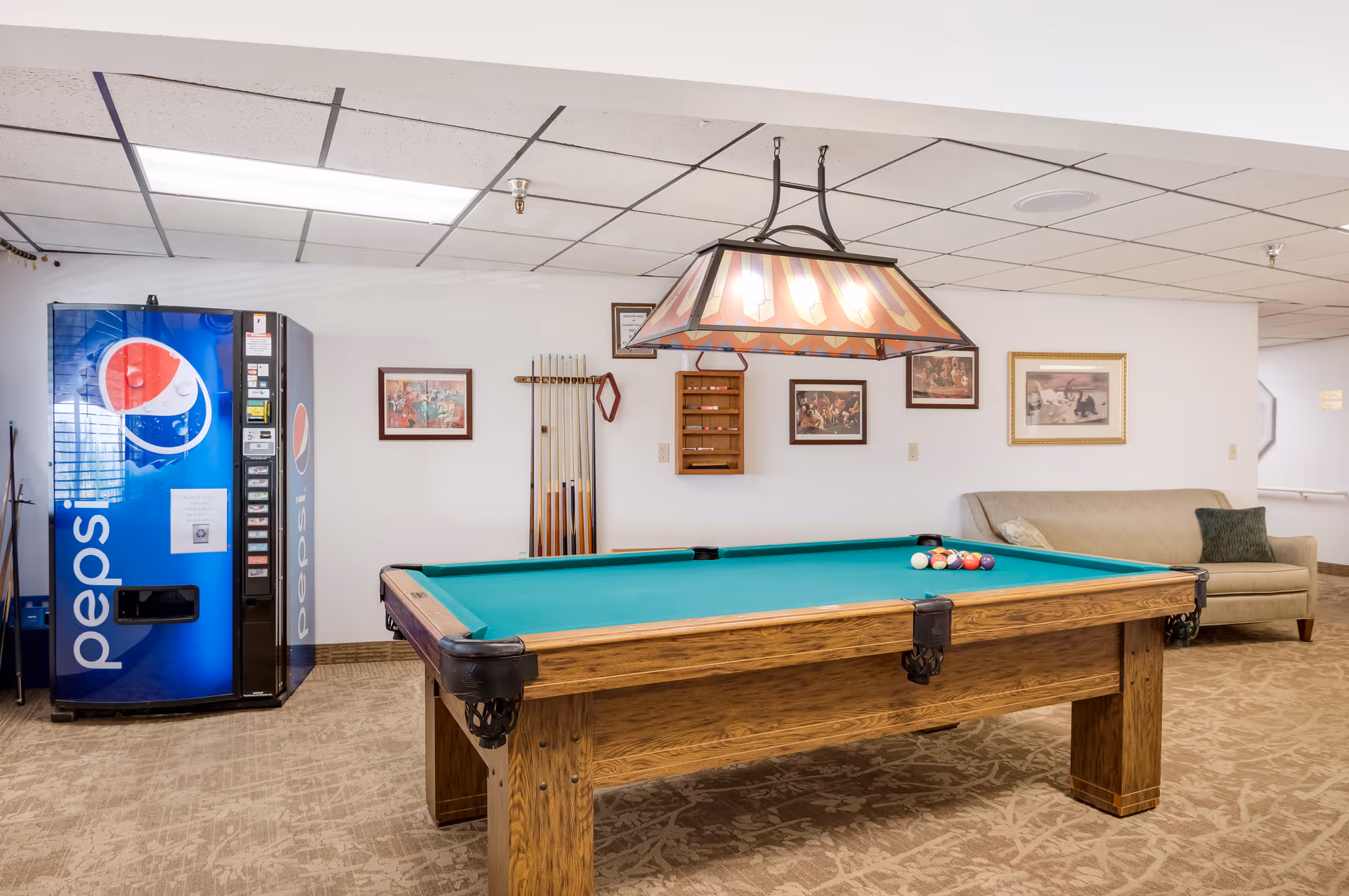 Pool table with hanging light, billiard cues, a Pepsi vending machine, and a sofa in a recreation room.