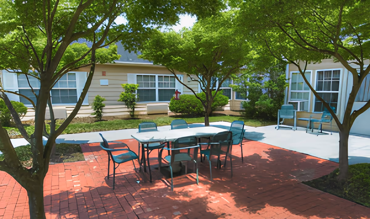 Shaded outdoor courtyard with a rectangular table and several chairs on a red brick patio in front of single-story building windows.