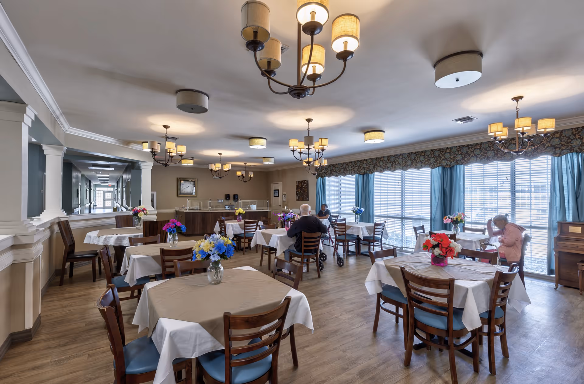 A bright dining room in a senior living facility with several tables covered in beige and white tablecloths, each adorned with colorful flower arrangements. There are wooden chairs with blue cushions around the tables. Large windows with blue curtains and floral valances let in natural light. Two elderly individuals are seated at separate tables near the windows. The room features multiple ceiling lights and chandeliers, and a hallway is visible through an open wall on the left side.