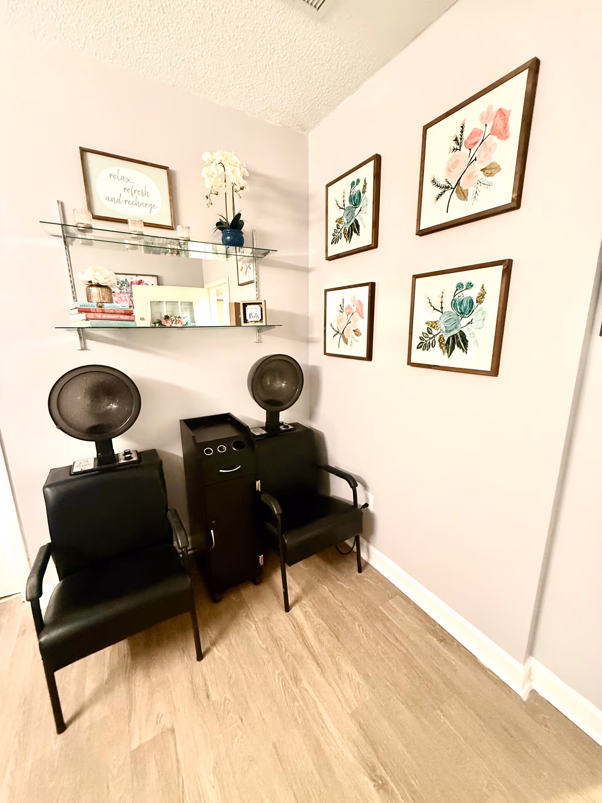 Corner of an interior salon area with two black hooded hair dryers, a small workstation, glass shelving with decor, and four framed floral prints on the wall.