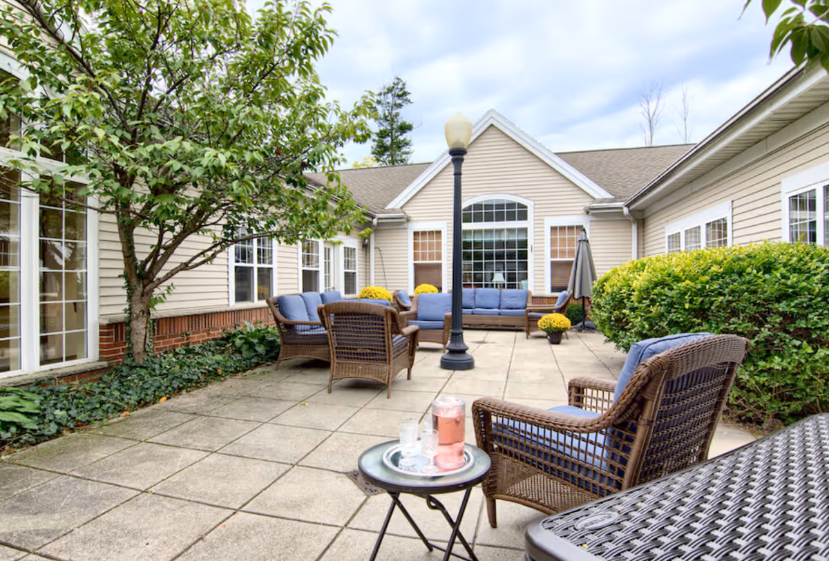 Outdoor patio area at Peregrine Senior Living at Orchard Park featuring wicker chairs with blue cushions arranged around a central lamp post, surrounded by greenery and beige building walls with multiple windows.