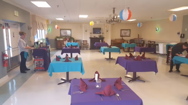 A spacious dining room with several tables covered in purple and teal tablecloths, each set with folded maroon napkins, utensils, and glasses. Two people are seen preparing the room, one on the left near a cart and the other on the right adjusting a table. The room is decorated with hanging beach balls and has a piano and framed pictures on the far wall.