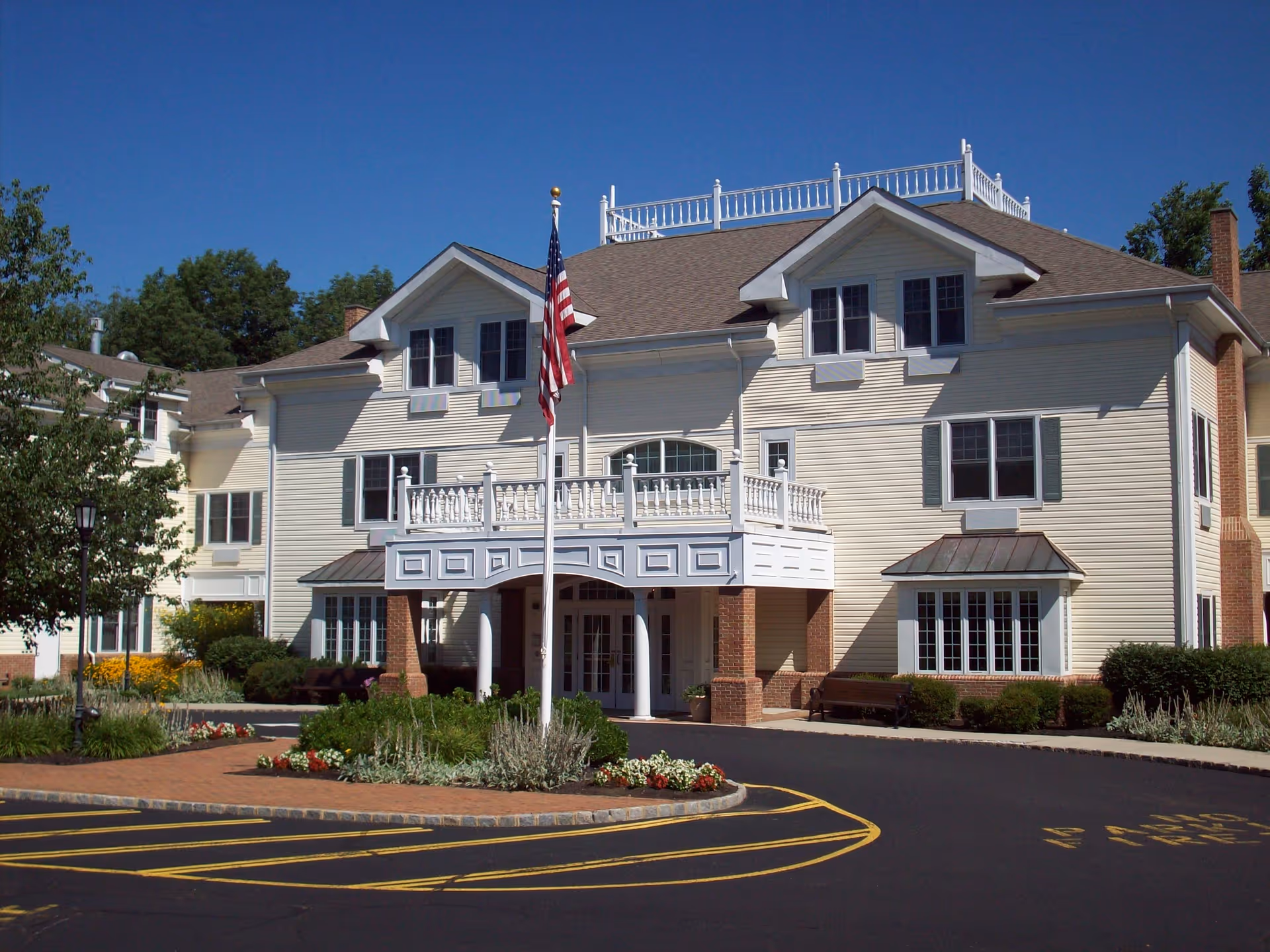 Front exterior view of a large, multi-story assisted living facility building with beige siding, white trim, and a brown roof. The entrance features a covered porch with white railings and columns, an American flag on a flagpole, landscaped flower beds, and a paved driveway with yellow markings.