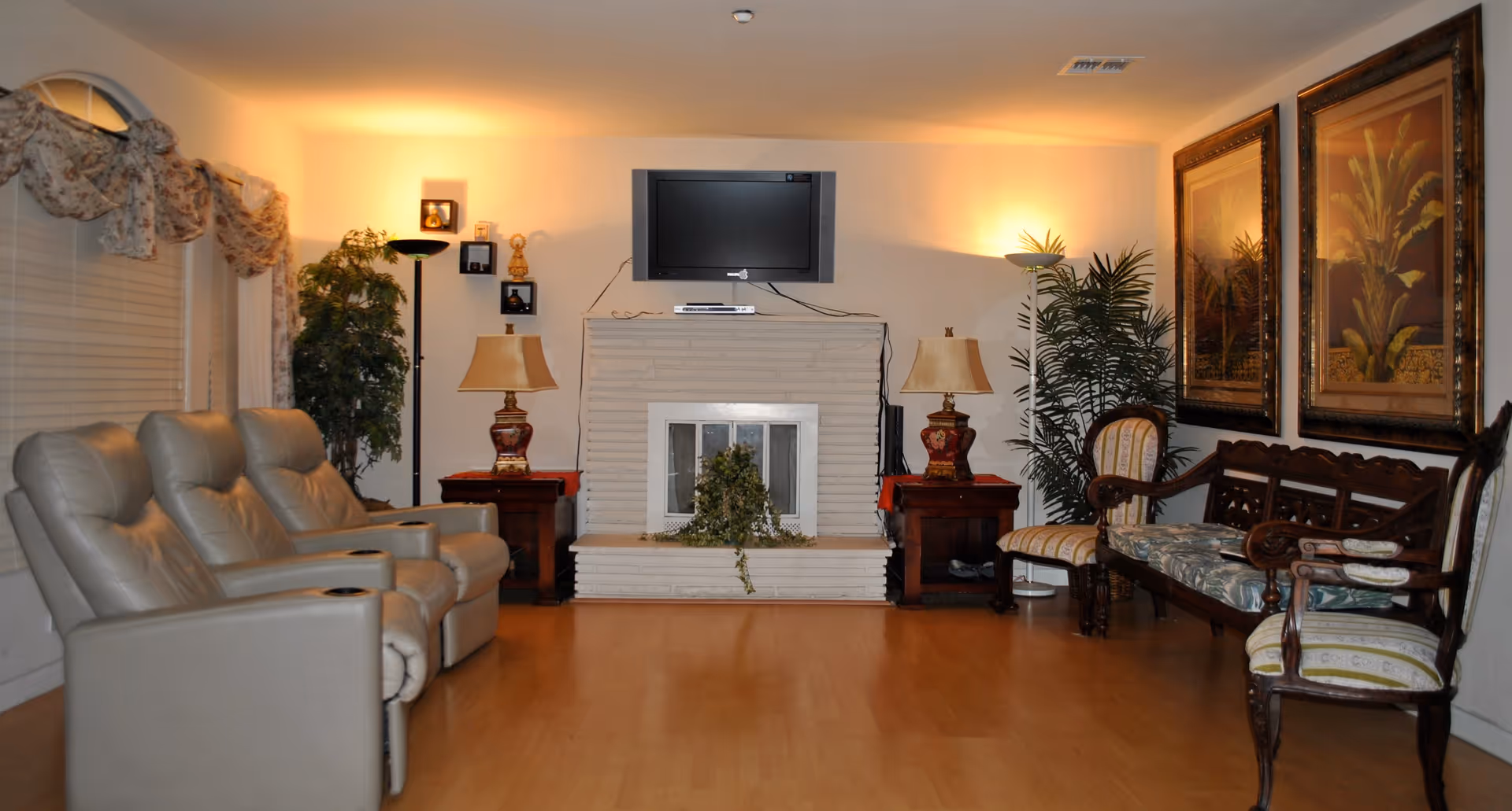A cozy living room with three beige recliner chairs on the left, a white brick fireplace in the center with a flat-screen TV mounted above it, two wooden side tables with lamps on either side of the fireplace, and a wooden bench with floral cushions and two matching chairs on the right. The room is warmly lit with wall sconces and floor lamps, decorated with framed botanical artwork and potted plants.