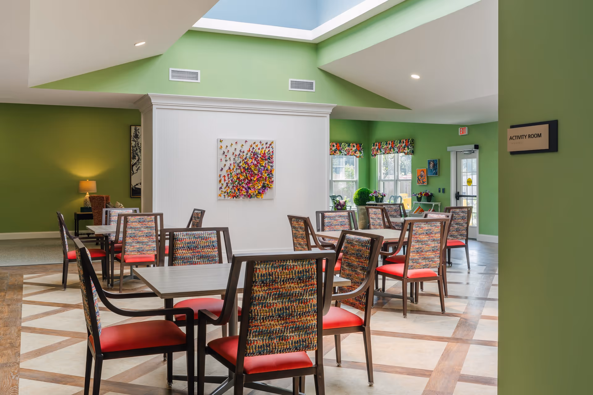 A brightly lit activity room with green walls and a skylight ceiling. The room contains several tables with chairs that have colorful patterned backs and red seats. There is a colorful butterfly wall art on a white partition, floral window valances, and various decorative plants and paintings near the windows. A sign on the wall reads 'ACTIVITY ROOM'.
