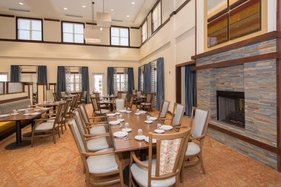 A spacious dining room with multiple wooden tables and chairs arranged neatly. Each table is set with white plates, glasses, and napkins. The room features large windows with blue curtains, high ceilings with additional windows near the top, and a stone fireplace on the right side. The carpet is patterned in warm tones, complementing the overall cozy and inviting atmosphere.