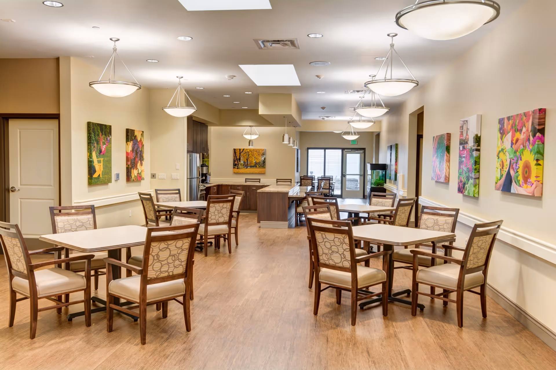 Bright communal dining room with multiple tables and chairs, pendant lights, artwork on the walls, and an open kitchen area at the back.