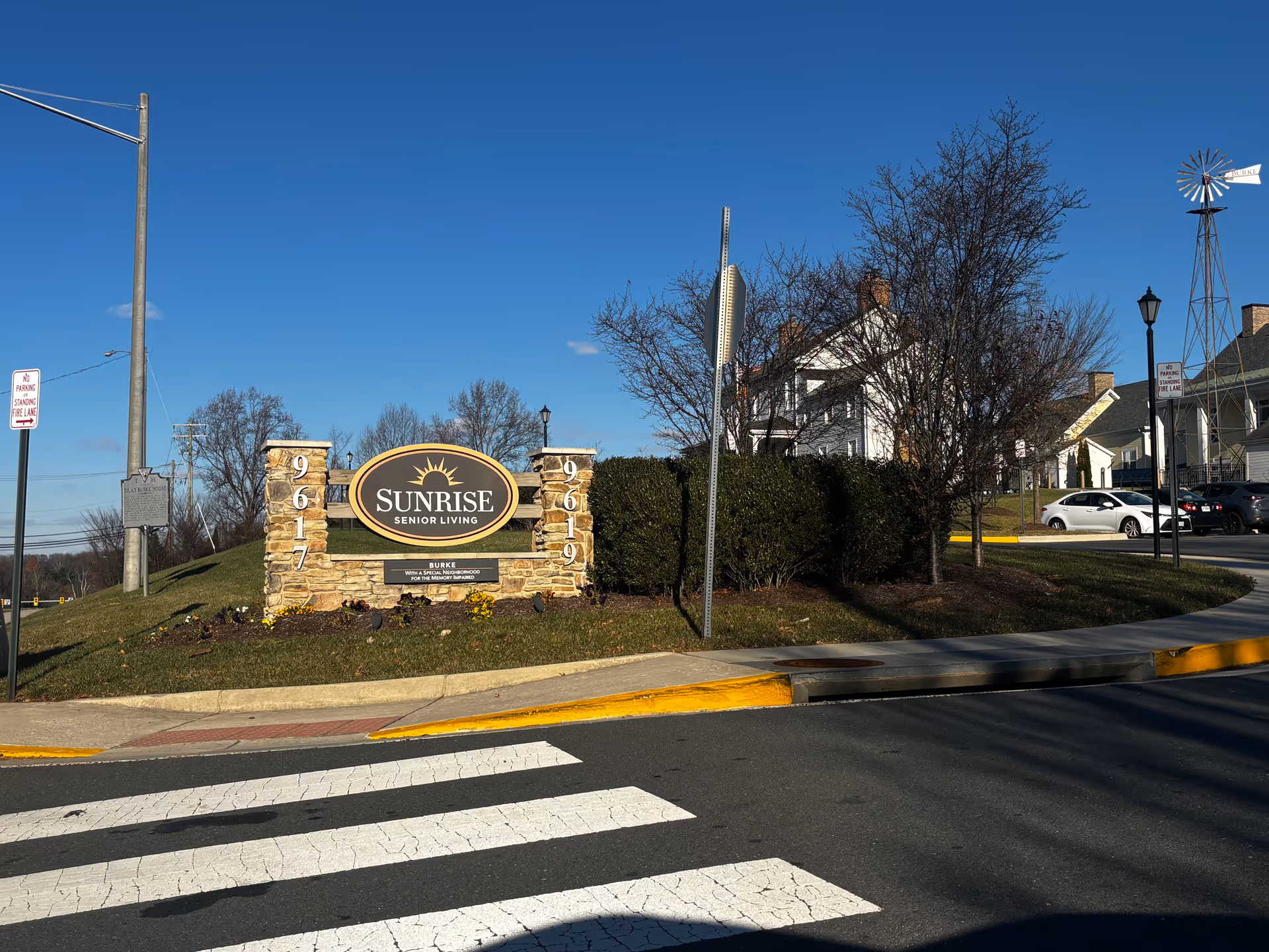 Stone entrance sign reading "Sunrise Senior Living" by a roadside crosswalk with the facility buildings and parked cars in the background.