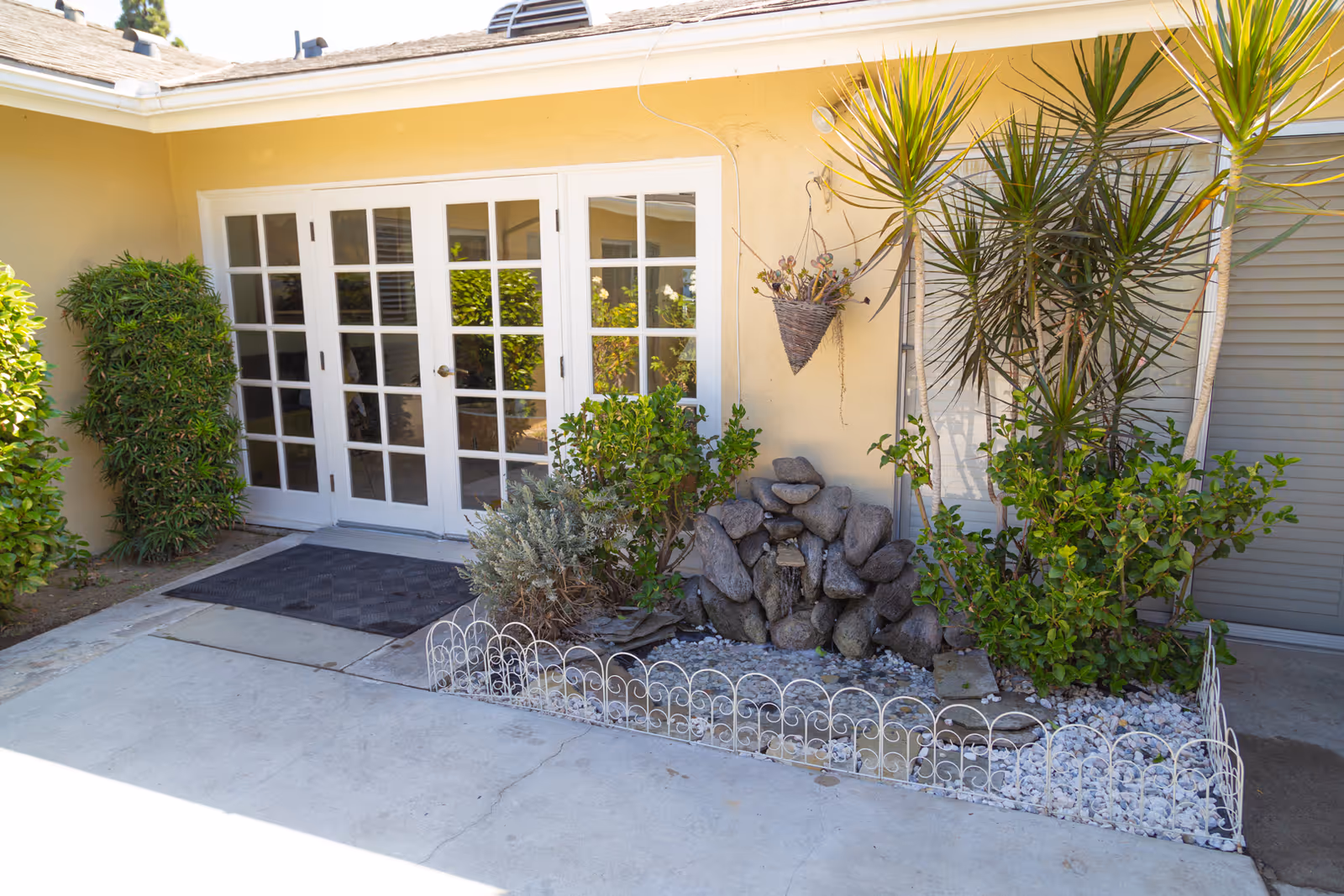 Outdoor patio area with a small garden bed containing rocks, plants, and a hanging basket in front of a building with beige walls and white-framed glass double doors.