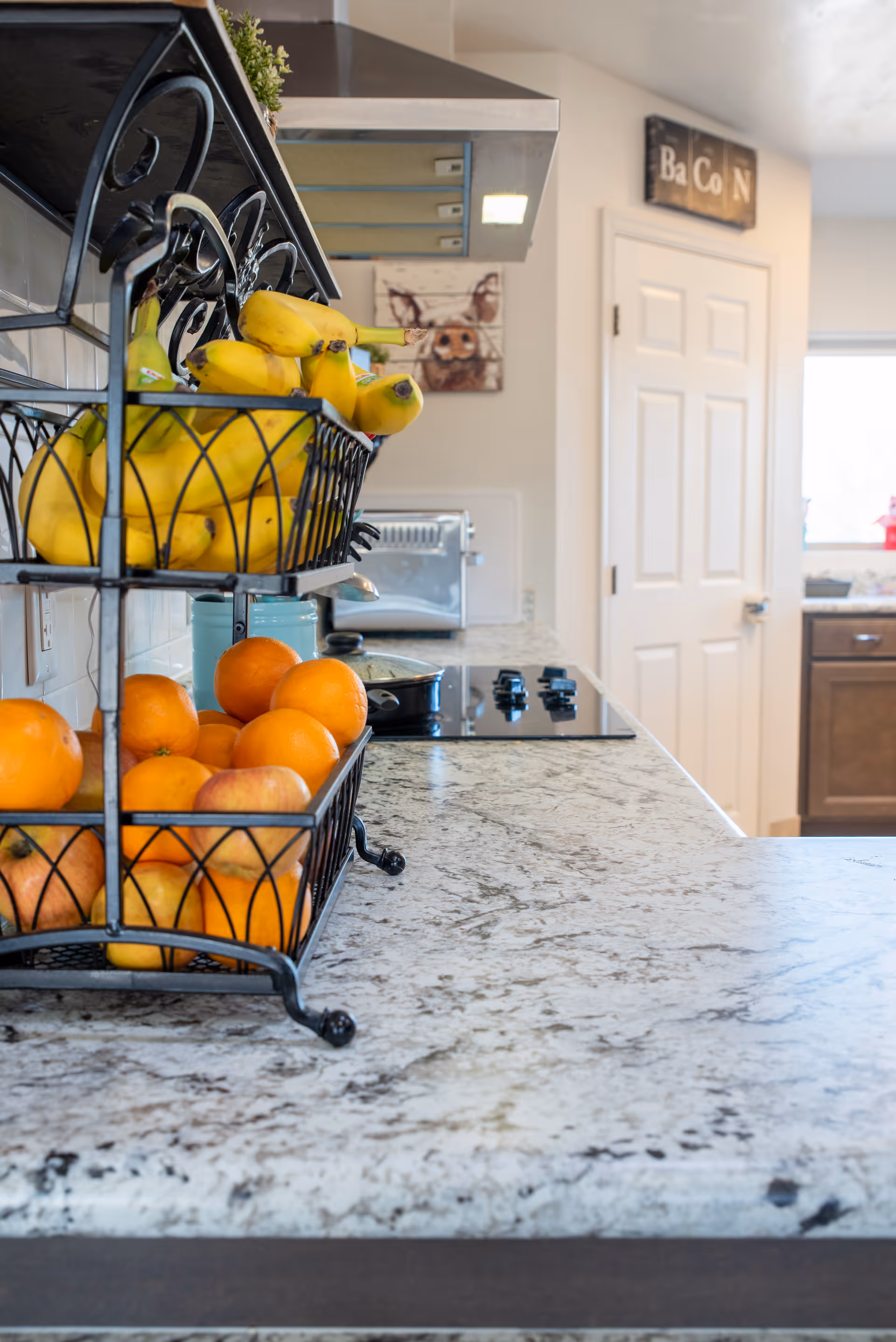 Close-up view of a kitchen countertop with a two-tiered black metal fruit basket holding bananas on the top tier and oranges and apples on the bottom tier. In the background, there is a stovetop with a pot, a toaster, a door, and a wall decoration with the word 'BaCoN'.