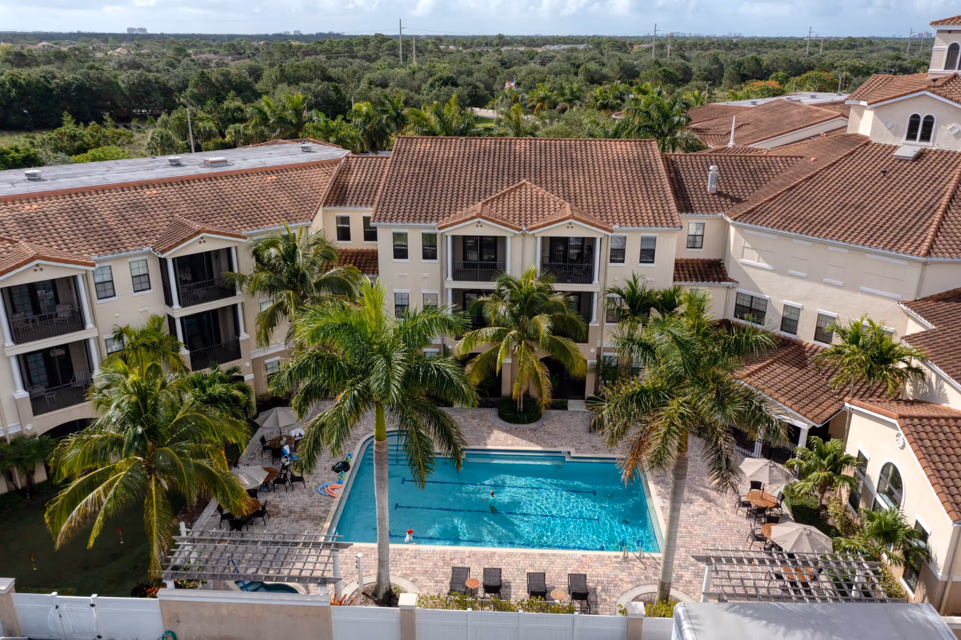 Aerial view of a senior living facility courtyard featuring a rectangular swimming pool surrounded by palm trees, lounge chairs, tables with umbrellas, and a multi-story building with terracotta roof tiles in the background.