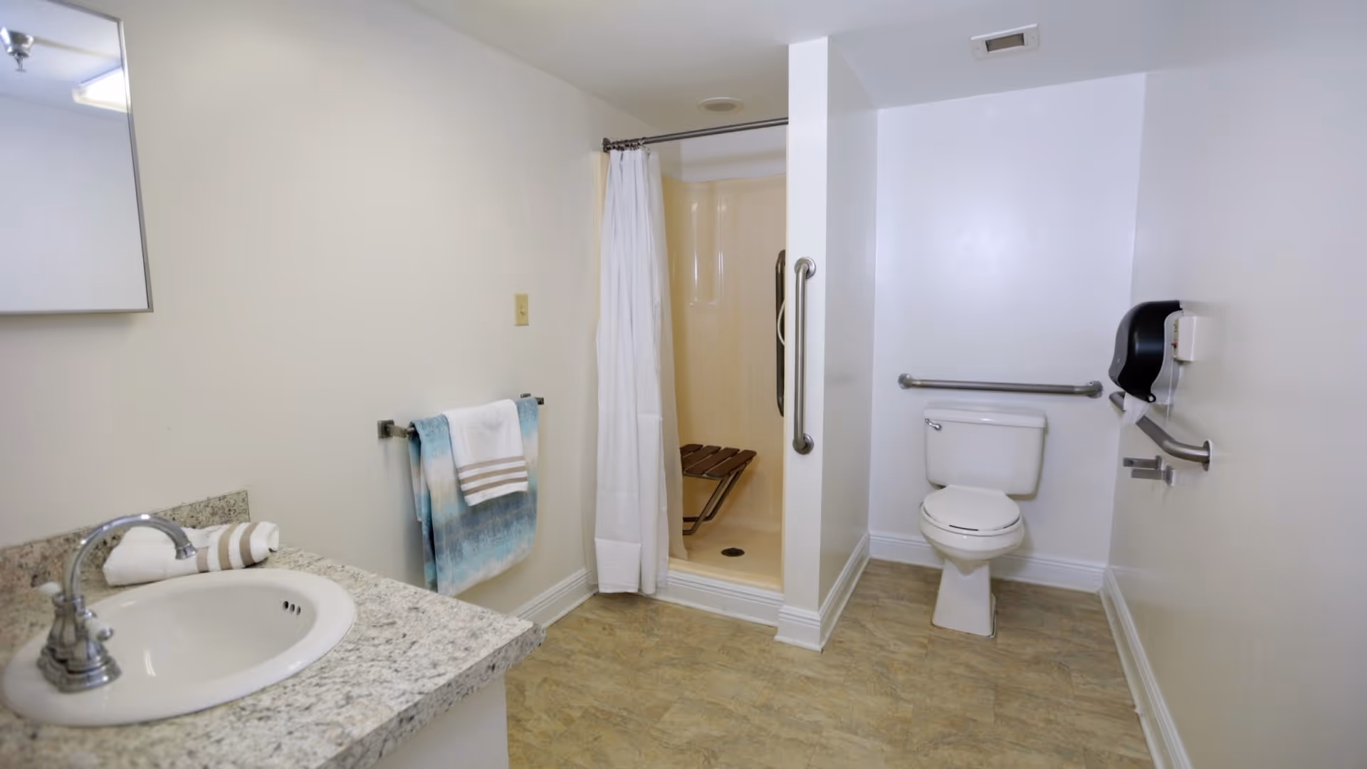 A clean and accessible bathroom featuring a white toilet with grab bars on the wall, a walk-in shower with a white curtain and a foldable wooden seat, a sink with a granite countertop, a mirror above the sink, and a towel rack with two towels hanging on it.