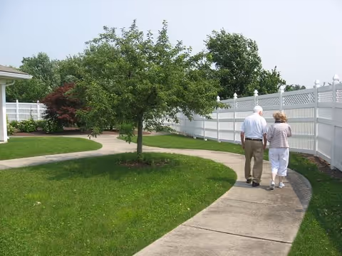 An elderly couple walking hand in hand on a curved concrete pathway in a green outdoor garden area with a white fence and trees surrounding them.