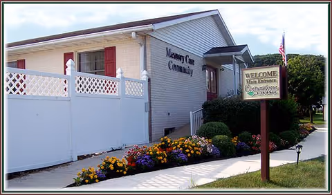 Exterior view of a single-story building with a white fence and colorful flower beds along the sidewalk. A sign near the walkway reads 'WELCOME Main Entrance Cornerstone Living'. The building has light-colored siding with red shutters and an American flag is visible in the background.