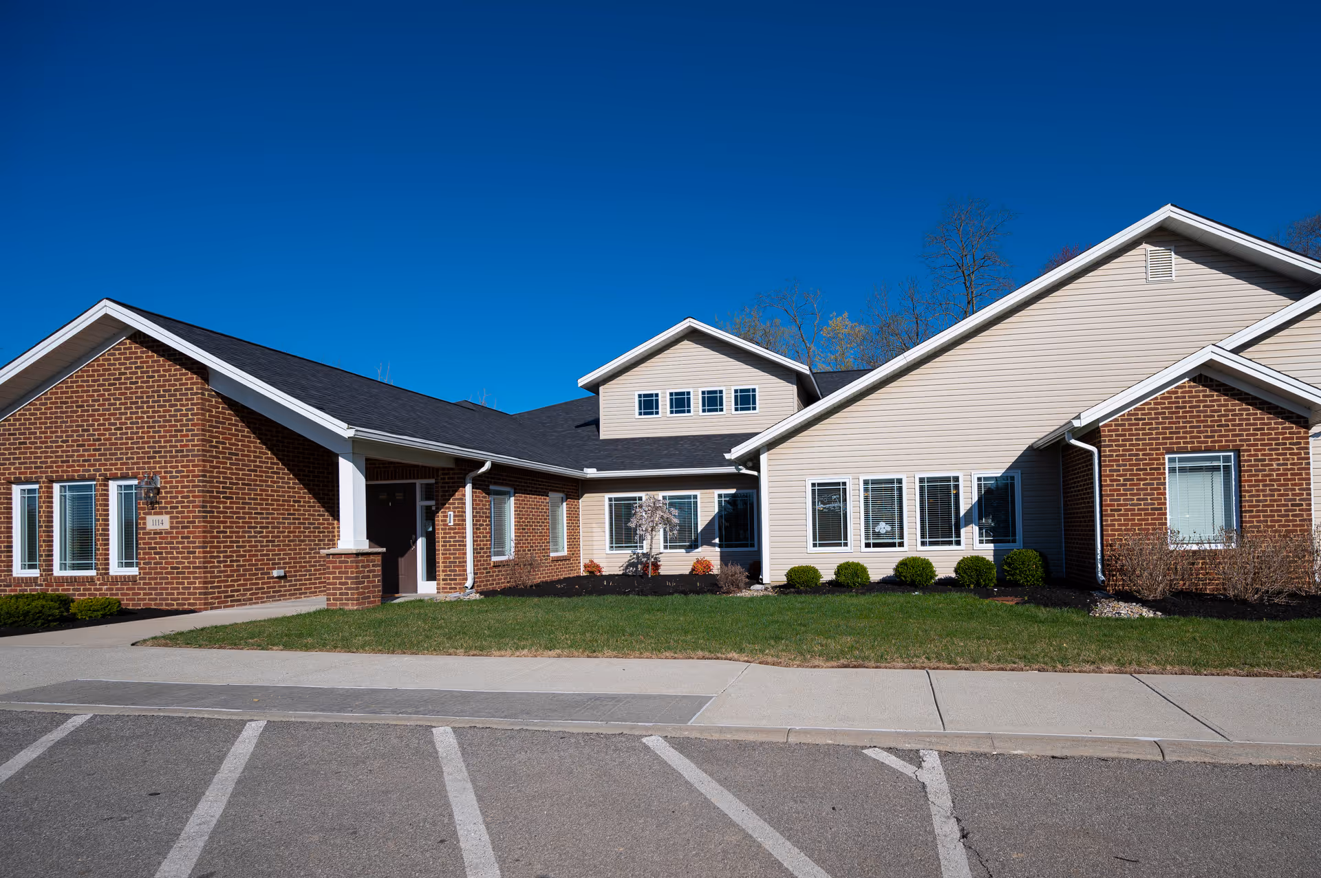 Single-story brick-and-siding senior living building with a front entrance, windows, small shrubs, and a clear blue sky.