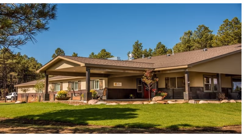 Single-story senior living home with a covered entryway, manicured lawn, and pine trees in the background.