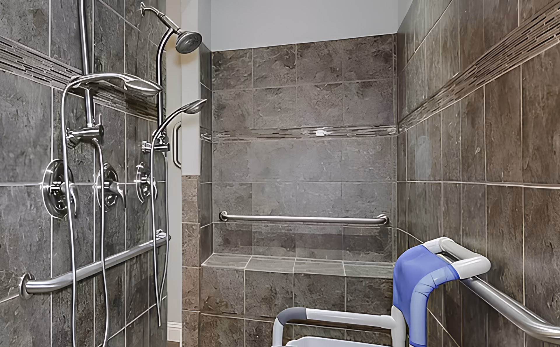 A tiled shower area with dark gray stone tiles, featuring a built-in bench, two chrome showerheads, and multiple grab bars. There is a blue and white shower chair positioned inside the shower for accessibility.