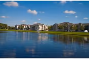 View of a residential apartment complex named Windsor Park Apartments across a large pond with clear blue sky and scattered clouds above.