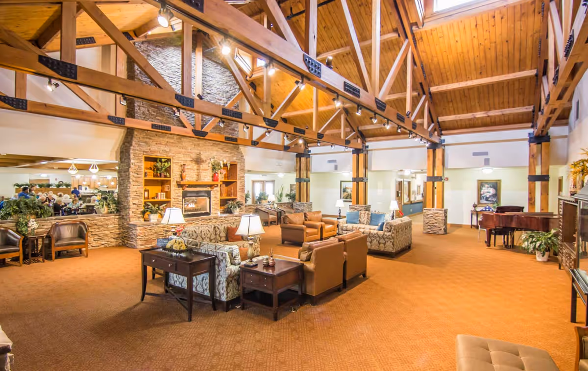 Spacious senior living facility common area with high wooden vaulted ceilings and exposed beams. The room features a large stone fireplace with built-in shelves, multiple seating arrangements including sofas and armchairs, side tables with lamps, and a grand piano in the corner. The carpet is a warm brown color, and the walls are light-colored with some artwork and plants placed around the room.