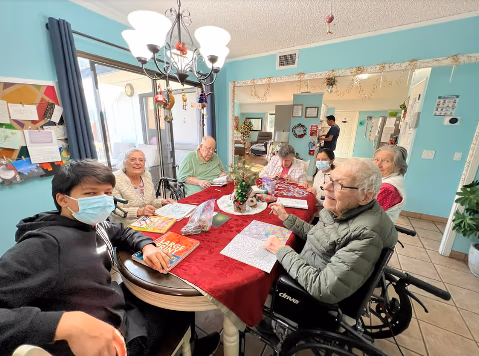 A group of elderly people and a younger person wearing a face mask sitting around a table with a red tablecloth, engaging in coloring activities. The room has light blue walls, a chandelier overhead, and a bulletin board on the wall. There are decorations hanging from the chandelier and a small Christmas tree centerpiece on the table. A man is seen standing in the background near a doorway.