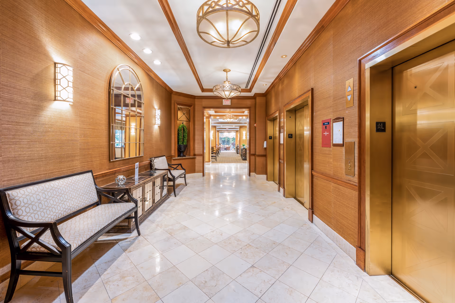 A well-lit hallway with three gold-colored elevators on the right side and a seating area with two benches and a cabinet on the left side. The walls are covered with a textured brown material, and there are decorative wall sconces and a large arched mirror above the cabinet. The floor is tiled with light-colored marble, and there are elegant ceiling light fixtures. The hallway leads to a more open area with additional seating and large windows in the background.