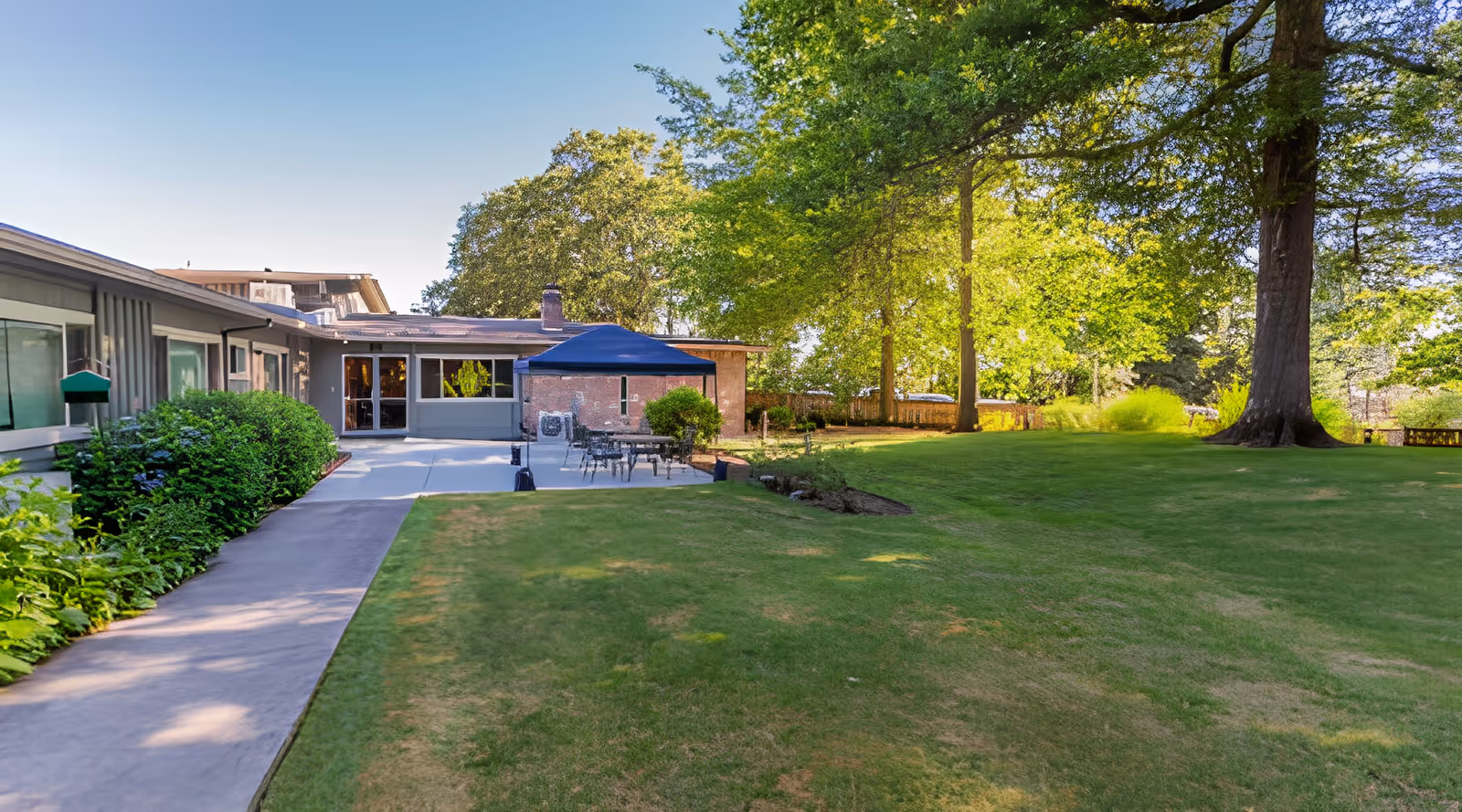 Outdoor view of Hillside Heights Rehabilitation Center showing a well-maintained lawn with large trees providing shade, a paved walkway along the building, and a patio area with tables and chairs under a blue canopy.