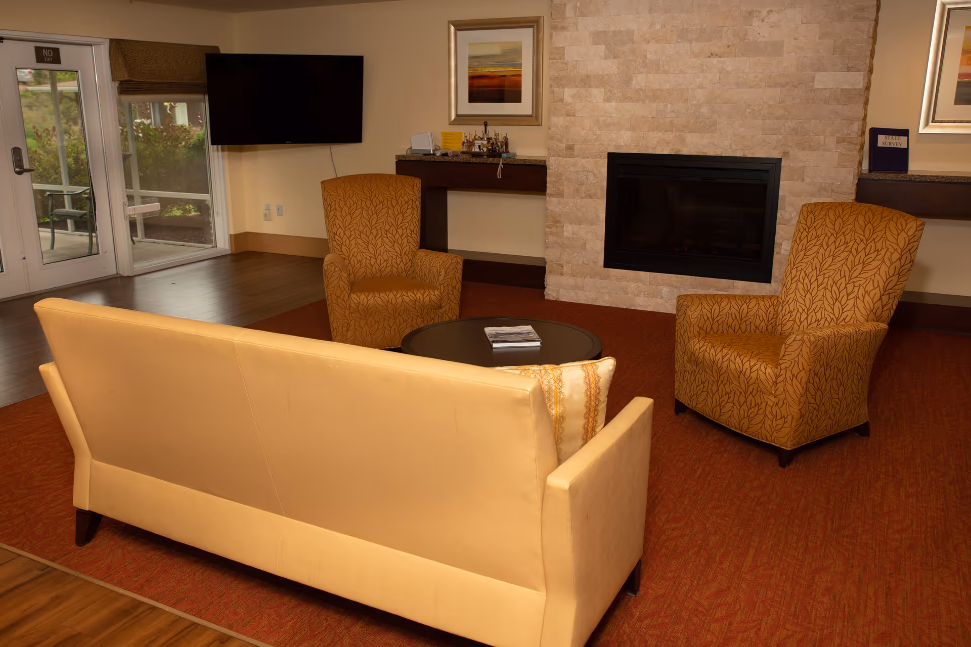 A cozy living room area with a beige sofa facing two patterned orange armchairs around a round black coffee table. Behind the seating area is a stone fireplace with a framed picture above it. To the left, there is a wall-mounted flat-screen TV and glass doors leading outside. The floor is a combination of wood and a red carpet.