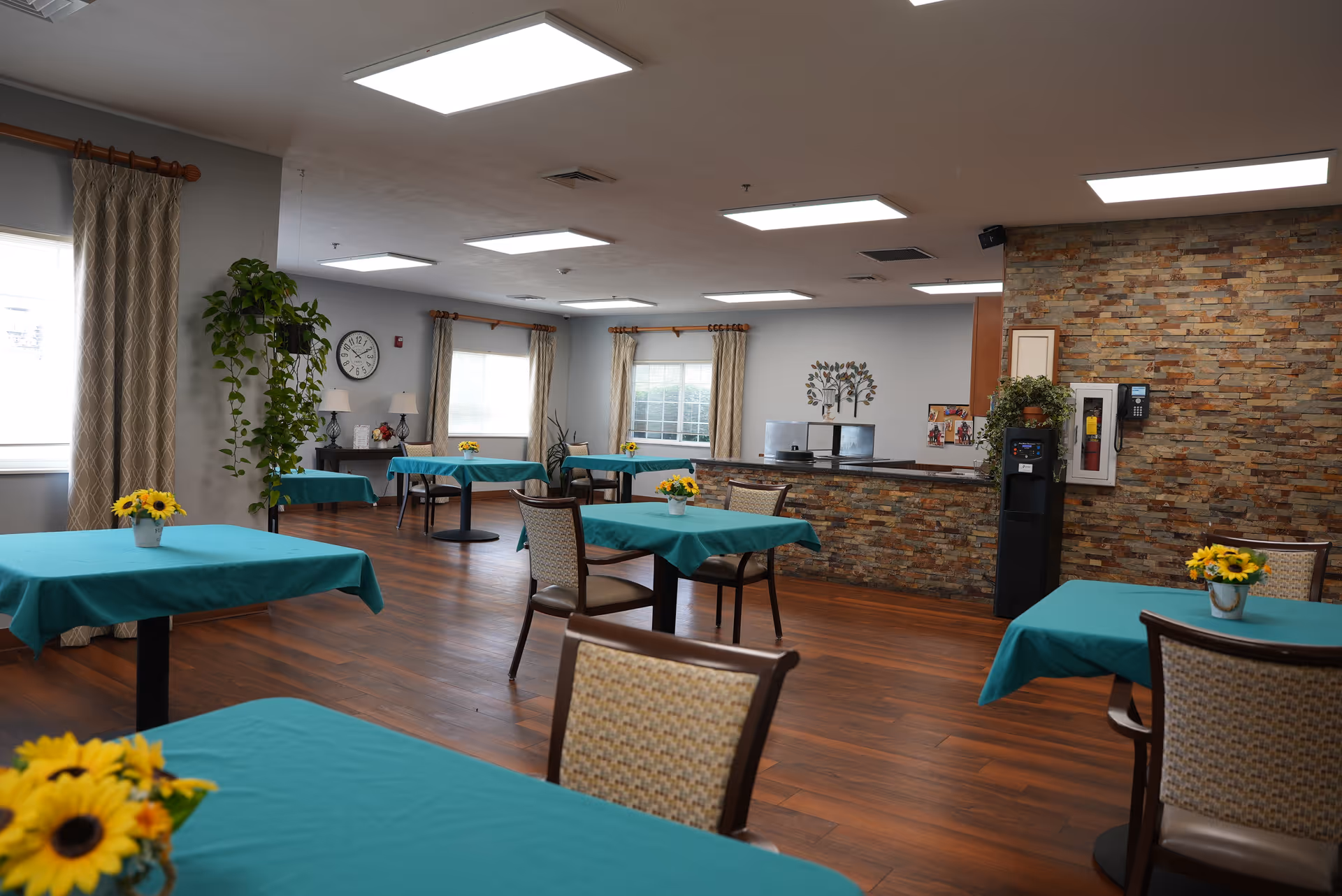 A spacious dining room with several tables covered in teal tablecloths, each adorned with a small vase of sunflowers. The room has wooden flooring, large windows with beige curtains, and a stone accent wall behind a counter area. There are chairs with woven backs around the tables, a water dispenser near the counter, and decorative plants and wall art in the background.