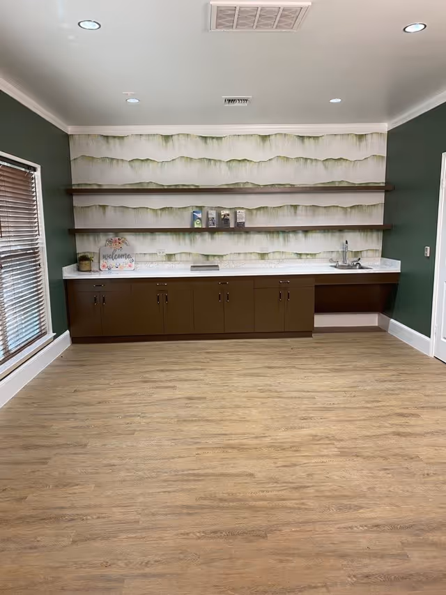 A room with green walls and light wood flooring featuring a long countertop with brown cabinets underneath, a small sink on the right, and three wooden floating shelves on the wall above. The back wall has a decorative wallpaper with a green and white pattern. A window with wooden blinds is on the left side of the room.