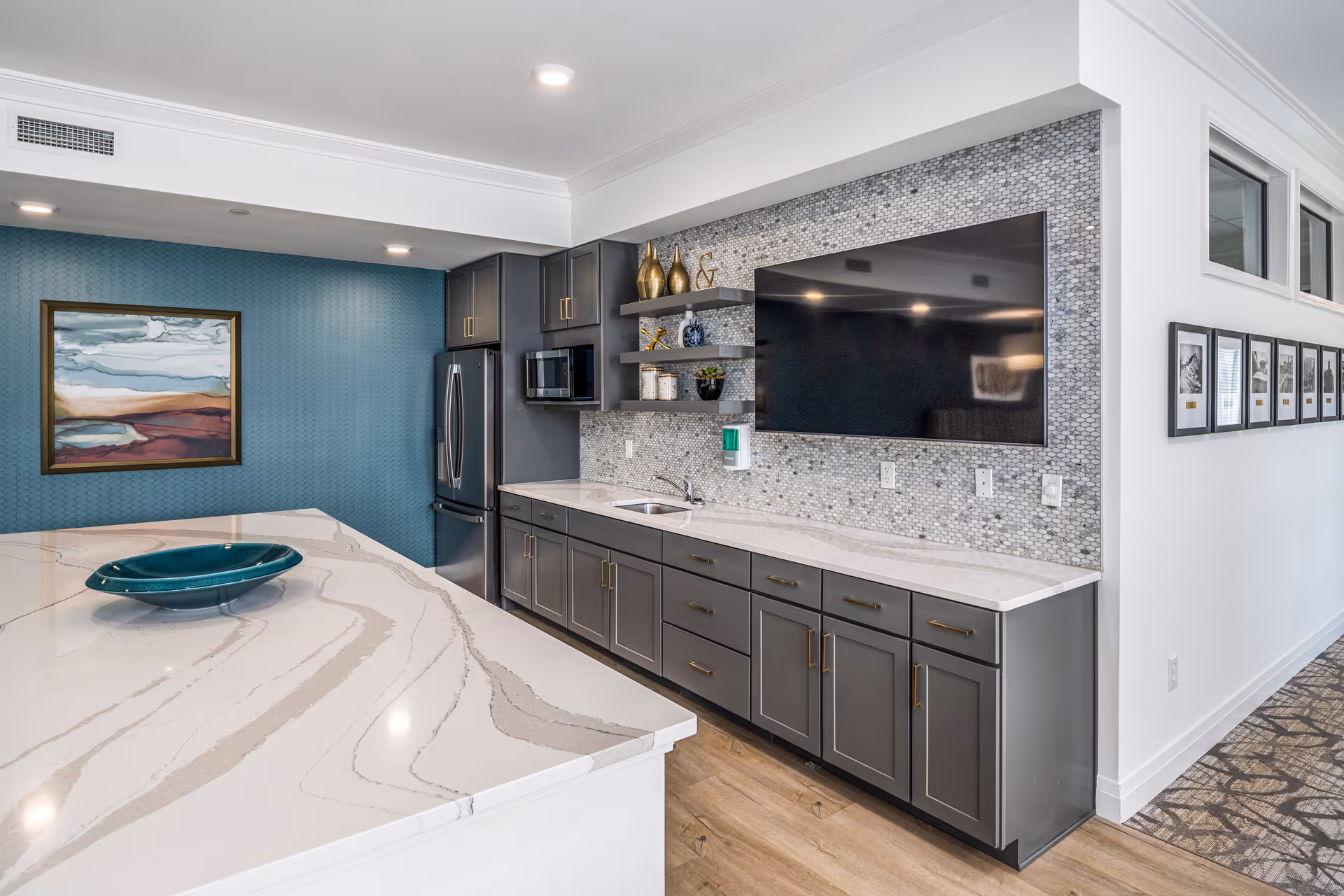 Modern communal kitchen with a large marble island, gray cabinets, stainless refrigerator, mosaic backsplash, and a wall-mounted TV.