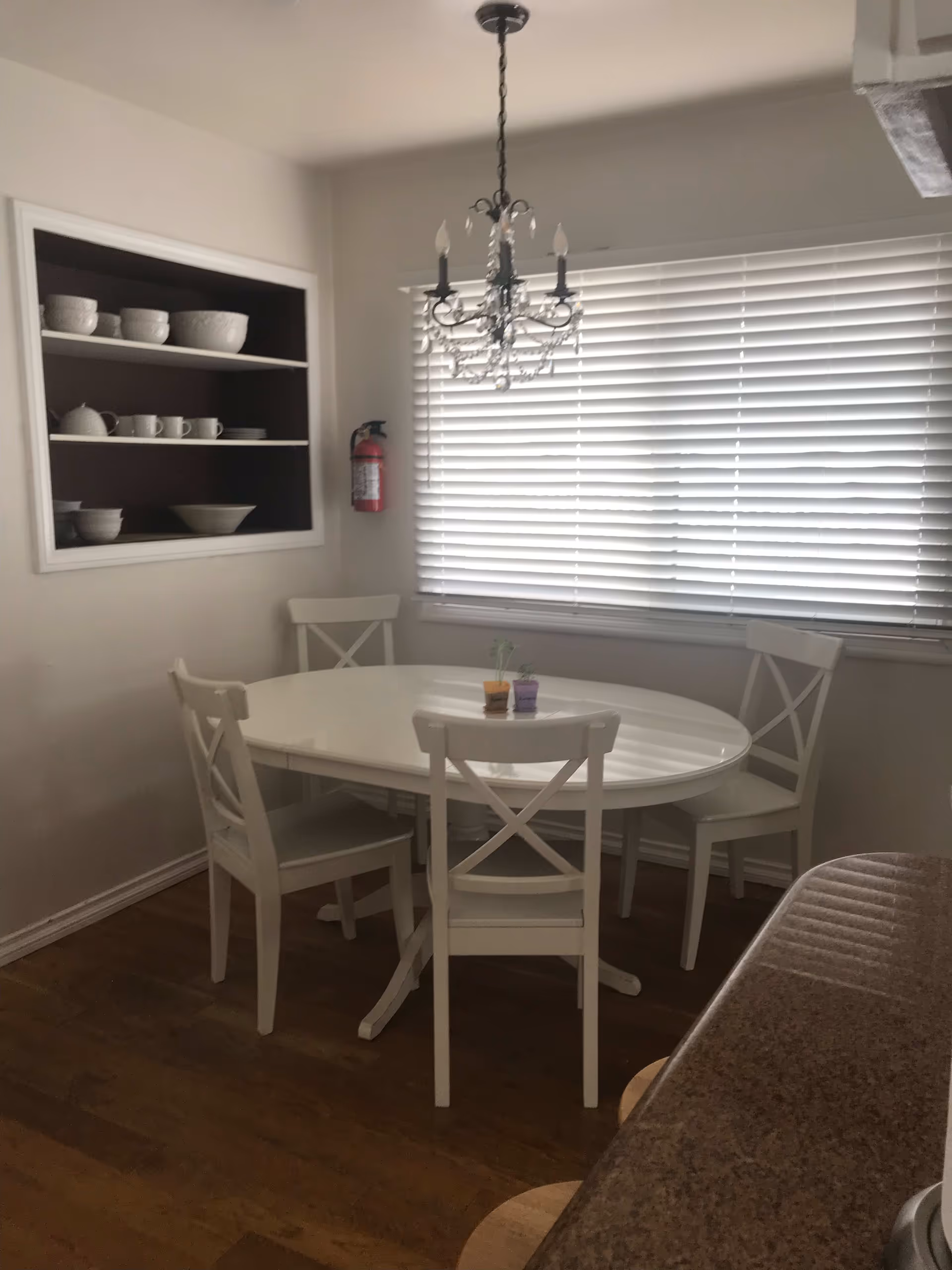 A dining area with a white oval table surrounded by four white chairs. Above the table hangs a decorative chandelier. There is a window with closed blinds behind the table, and a built-in shelf on the wall holding white bowls, cups, and plates. A fire extinguisher is mounted on the wall next to the shelf. The floor is wooden, and part of a kitchen counter with stools is visible on the right side.