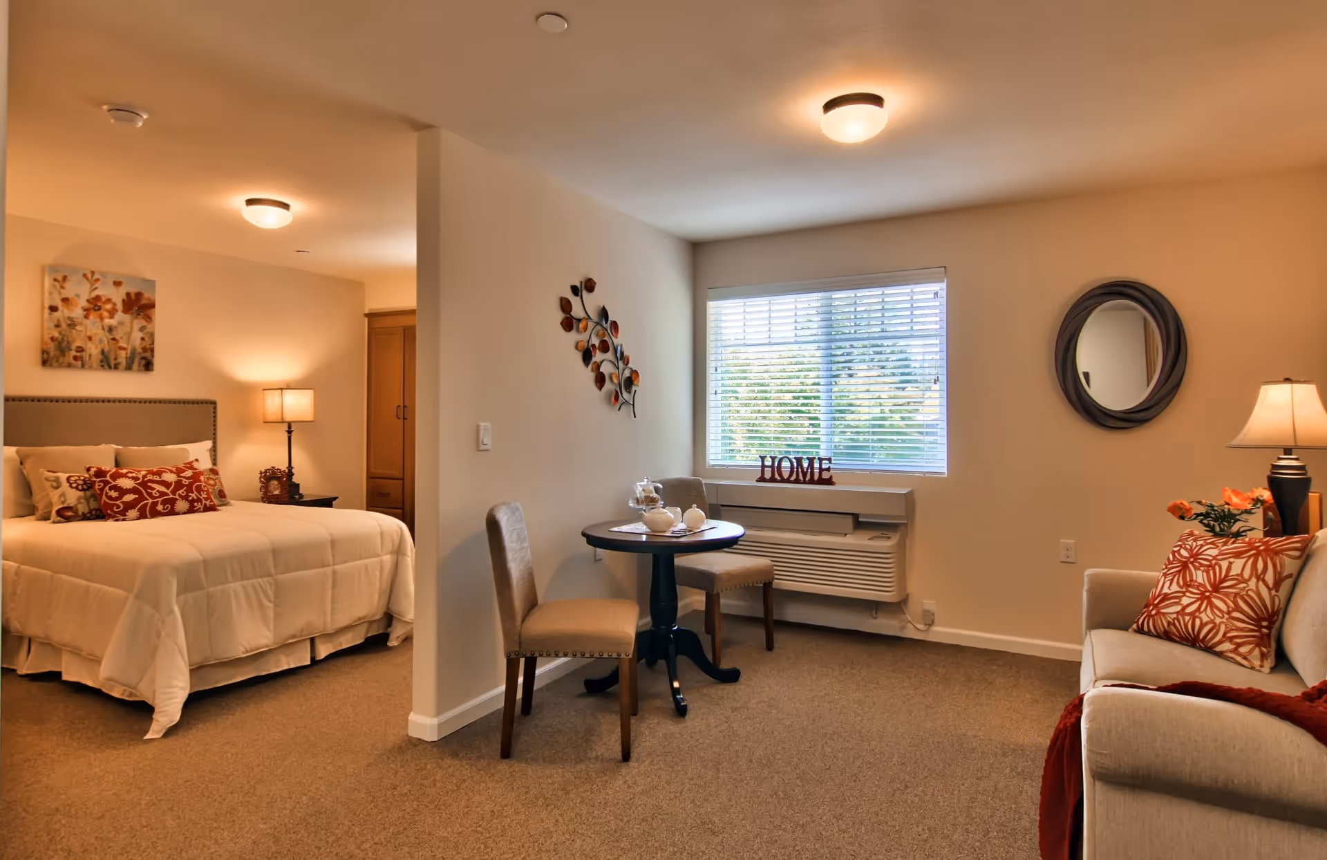 A cozy senior living apartment featuring a bedroom with a neatly made bed adorned with decorative pillows and a bedside lamp. Adjacent to the bedroom is a small dining area with a round table and two chairs, set near a window with blinds and a decorative 'HOME' sign on the windowsill. A comfortable sofa with patterned cushions and a floor lamp are also visible in the living area, along with wall decorations including a round mirror and a metal leaf art piece.